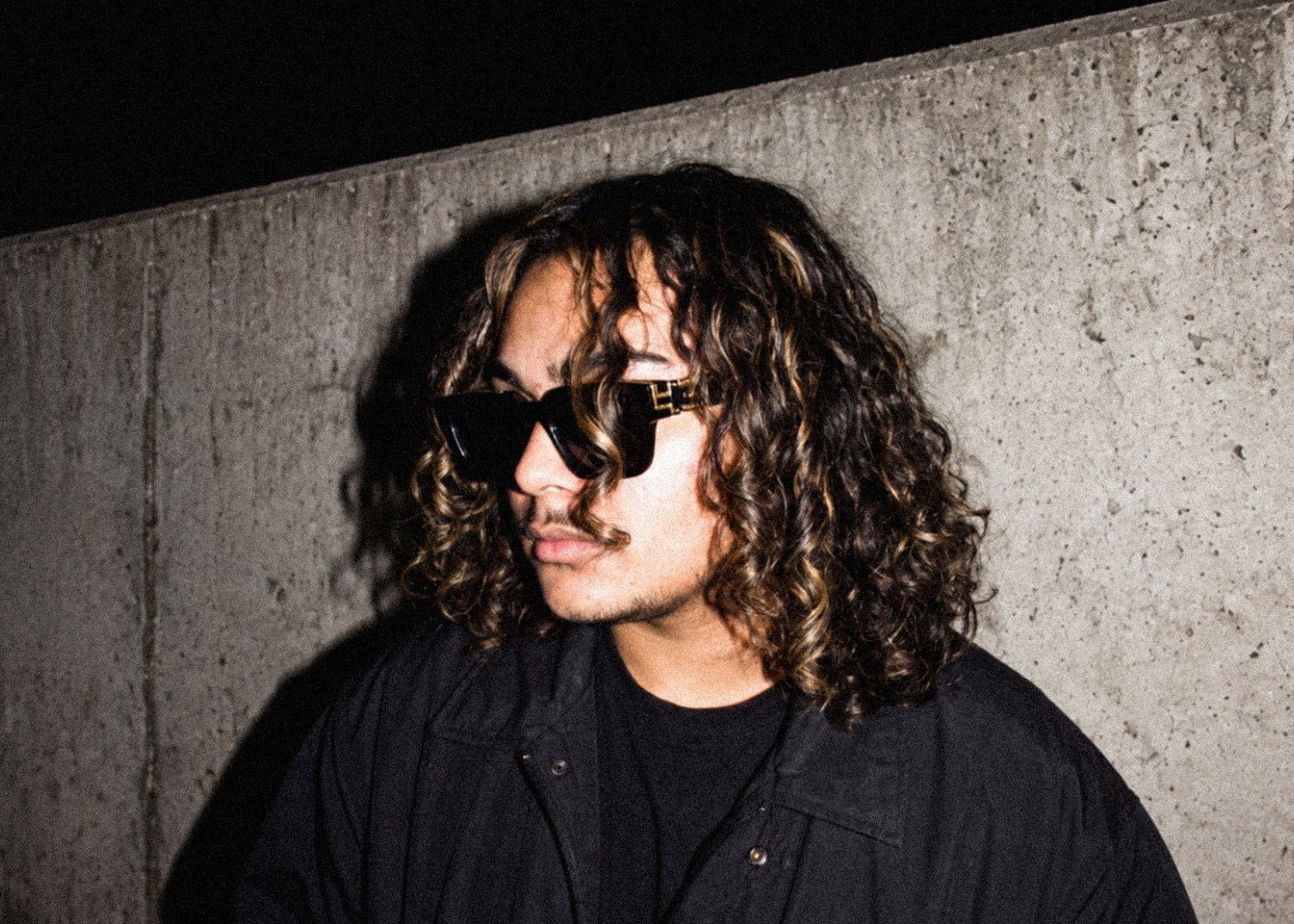 PRXD. JAY with curls and square sunglasses against the flash-lit concrete wall.