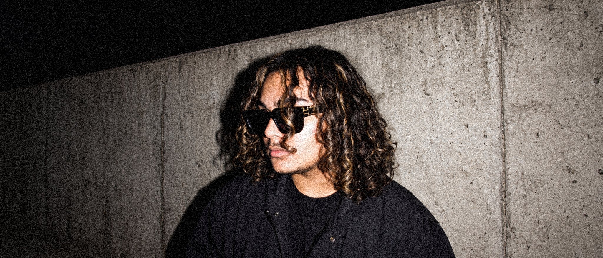 PRXD. JAY in square sunglasses and a black jacket, lit by camera flash against a concrete wall at night.